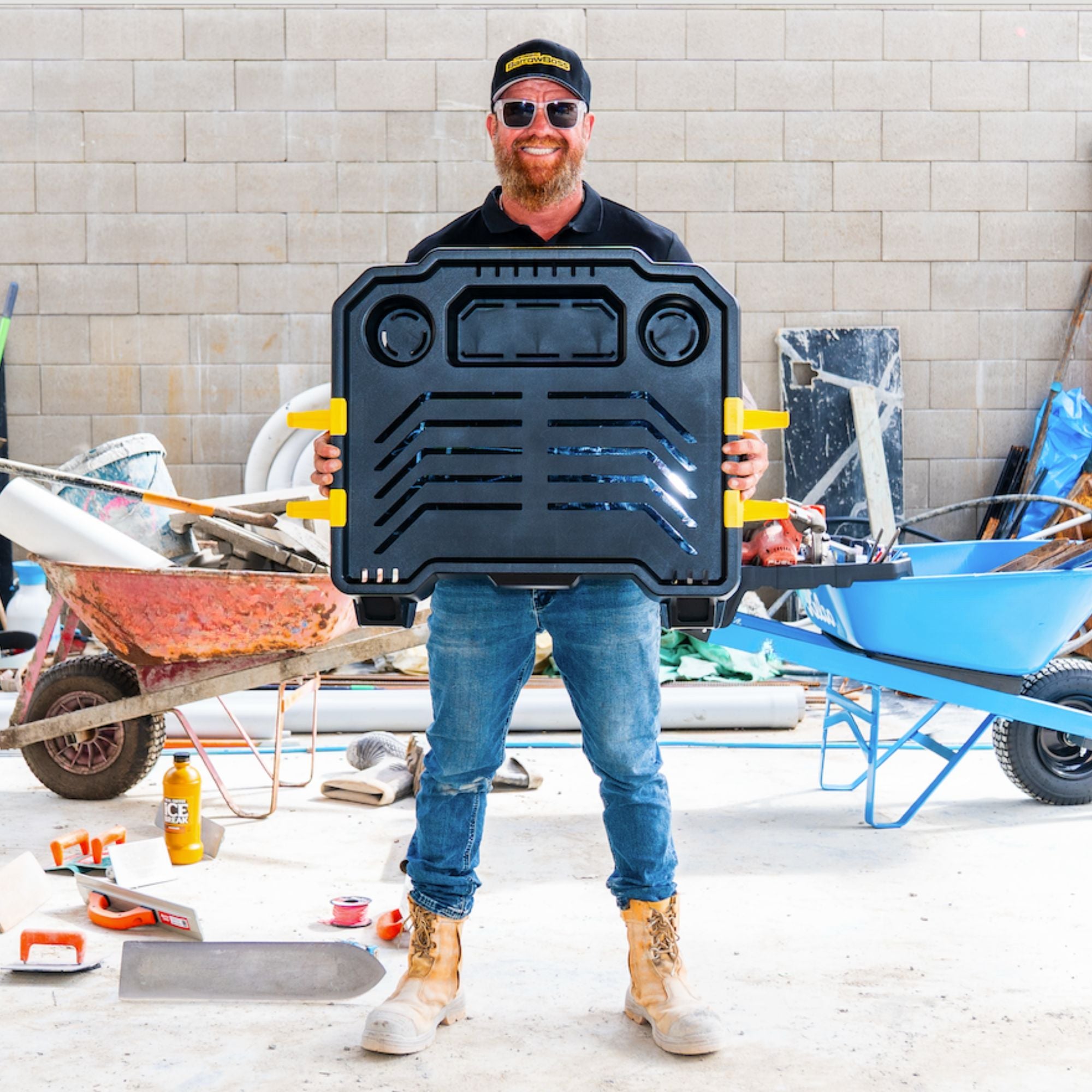 Tradesman holding the Barrow Boss wheelbarrow workstation attachment between a standard wheelbarrow and a Barrow Boss-equipped wheelbarrow at a construction site.