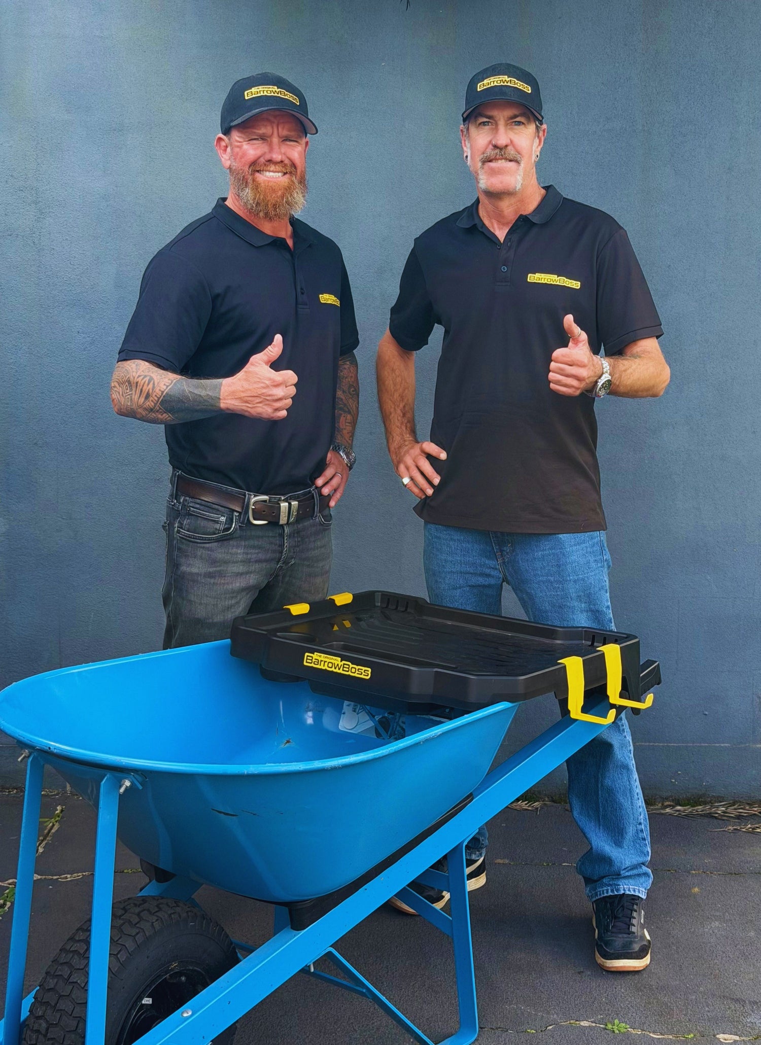 Barrow Boss founders Andre and Scott standing beside a wheelbarrow fitted with the Barrow Boss workstation attachment.
