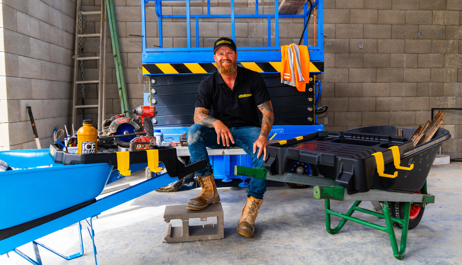 Andre, founder of Barrow Boss, sitting beside the Barrow Boss wheelbarrow workstation attachment on a construction site.